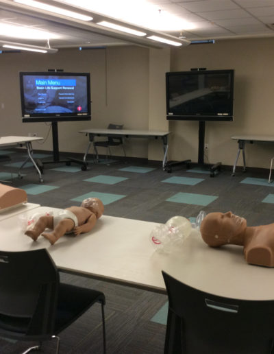 Image of a BLS classroom set-up with manikins, tables, TV monitors and other training gear.